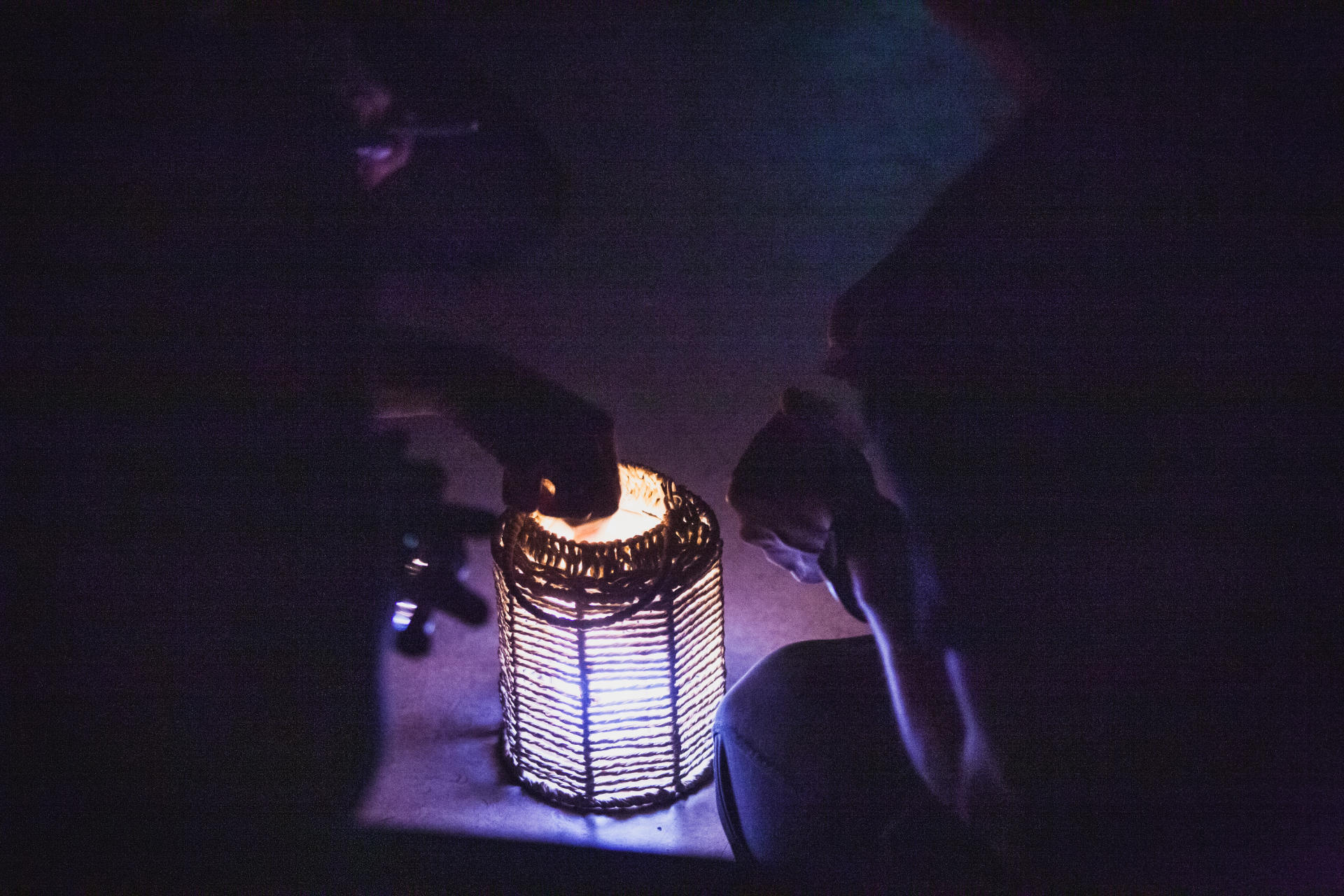 Silhouette of three people in a dark room, one of them is reaching into a lit latern that stands between them.